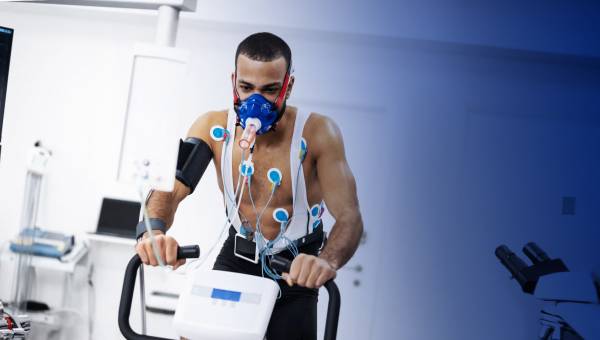 Ein afroamerikanischer Sportler führt mit freiem Oberkörper, verkabelt und mit medizinischer Sauerstoffmaske Tests auf einem Cardiofahrrad in der Praxis durch