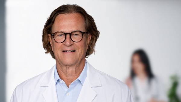 Ein Artz, Dr. Wilhelm Knarr, steht in einem hellen Praxisraum und lächelt freundlich
